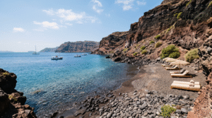 wide coastal scene of a secluded volcanic beach in Santorini, Greece, unusual dark volcanic sand and smooth black pebbles meeting crystal-clear blue water,