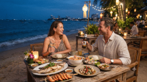 beachfront seafood dinner in Pattaya, Thailand, adult couple enjoying a table filled with grilled prawns, fresh fish, Thai seafood dishes and tropical drinks,