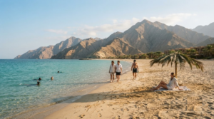 professional travel photograph of Al Aqah Beach, Fujairah, UAE, wide sandy beach meeting crystal-clear turquoise Gulf of Oman water, dramatic Hajar Mountains rising behind the coastline