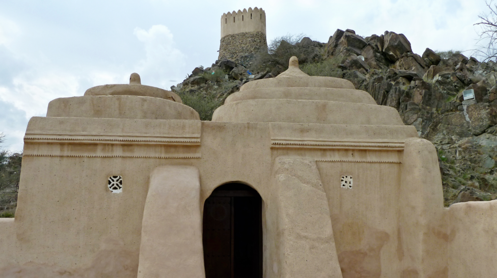 professional architectural travel photograph of Al Bidya Mosque, Fujairah, UAE, authentic small historic mosque with four distinctive rounded domes, traditional mud and stone construction,