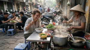 professional culinary travel photograph at a busy local street-food area in Vietnam, real adult international traveller sitting on a small low stool enjoying a bowl of authentic pho
