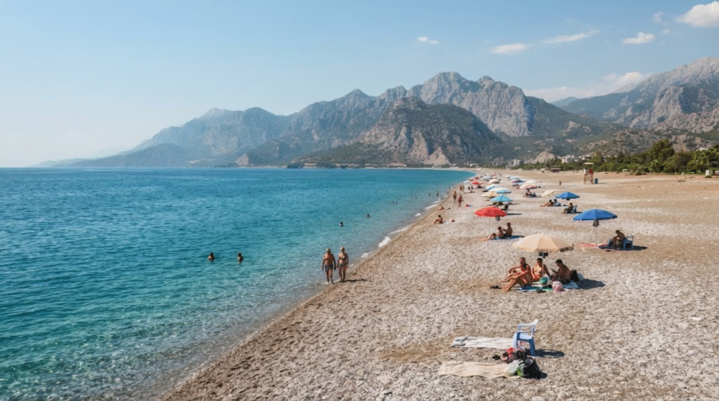wide-angle beach photograph of Konyaalti Beach in Antalya, Turkey, long pebble shoreline beside crystal-clear deep turquoise Mediterranean water,