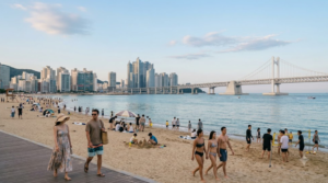 photograph of tourists relaxing and walking along Haeundae Beach, Busan, South Korea, blue ocean, sandy shoreline and modern skyline