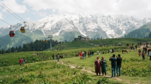 travel photograph of Gulmarg, Kashmir, featuring vast green mountain meadows transitioning into snow-covered Himalayan slopes, distant peaks and the Gulmarg Gondola visible naturally in the landscape.